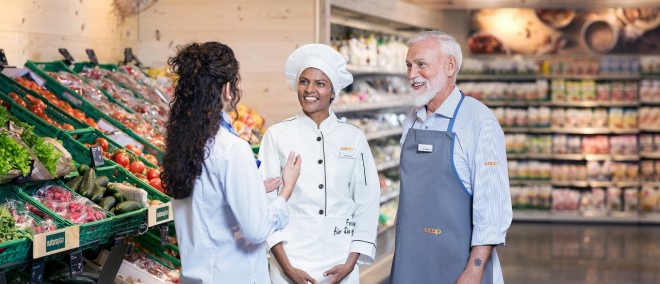 Eine Verkäuferin, eine Köchin und ein Metzger unterhalten sich in der Obst- und Gemüseabteilung des Coop Supermarkts.
