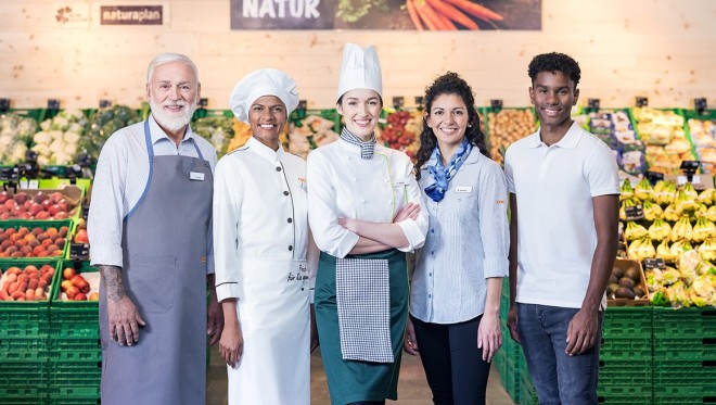 Eine Gruppe von Coop Mitarbeitenden lächelt vor der Obst- und Gemüseabteilung des Coop-Supermarktes in die Kamera.